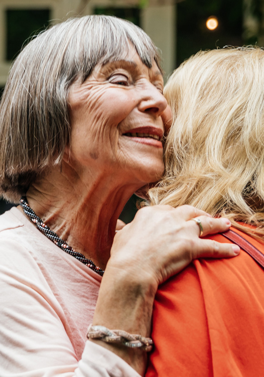 An older woman smiles as she embraces a friend at an outdoor family gathering.